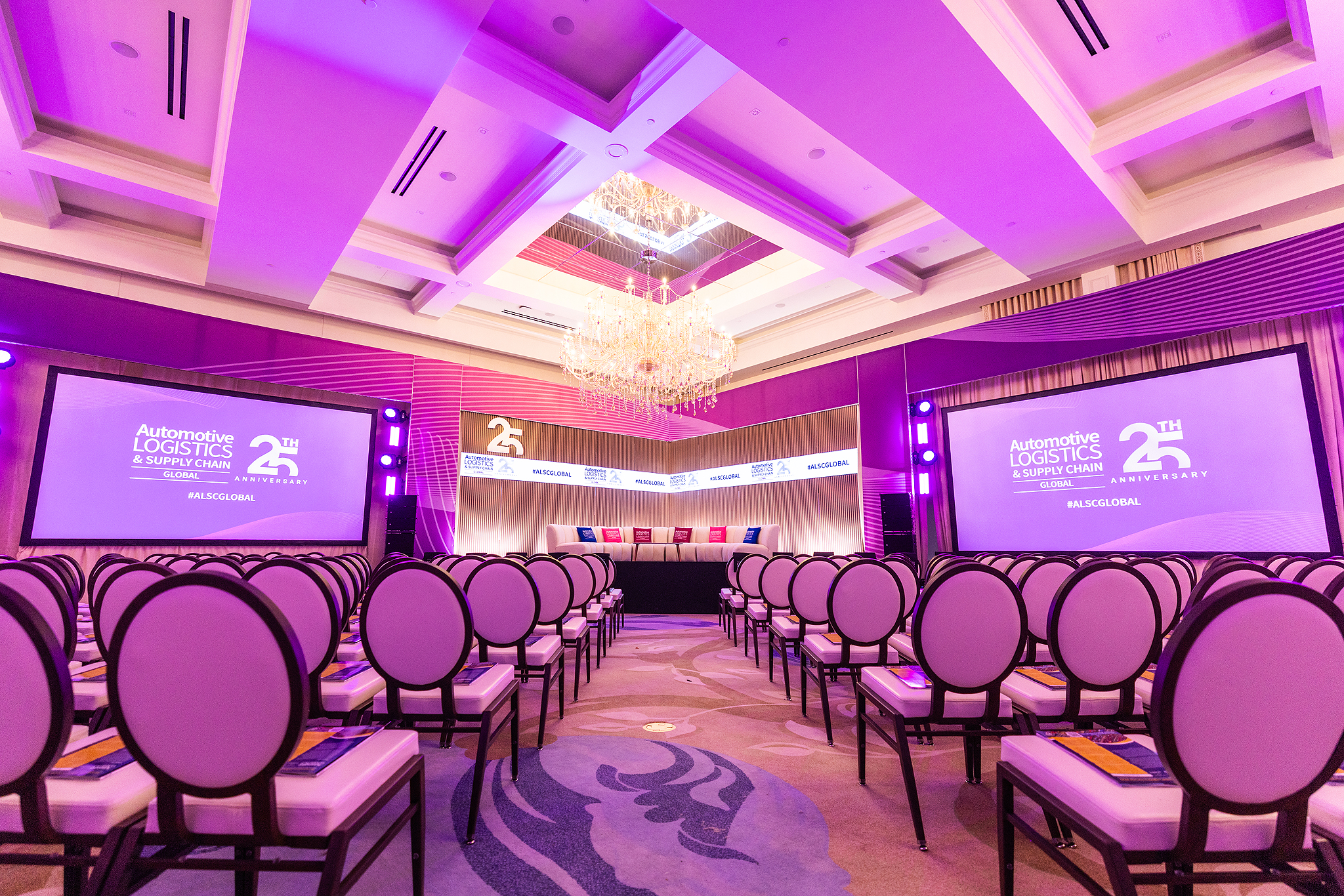 A brightly lit conference room at St Johns Resort in Detroit features rows of empty chairs facing a stage with a 25th anniversary Automotive Logistics and Supply Chain Global banner and two large projection screens, all accented by purple lighting.