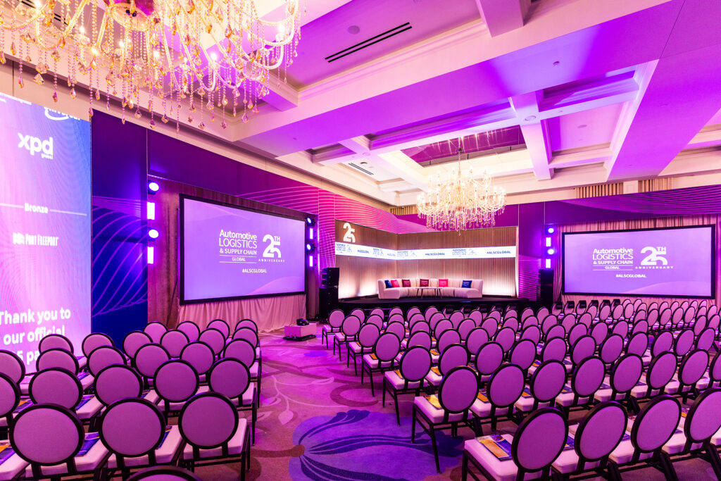 A brightly-lit corporate conference room at St Johns Resort in Detroit features rows of empty chairs facing a stage, two large screens, and purple lighting. Chandeliers hang from the ceiling as the stage awaits the “Automate Logistics Summit.”.