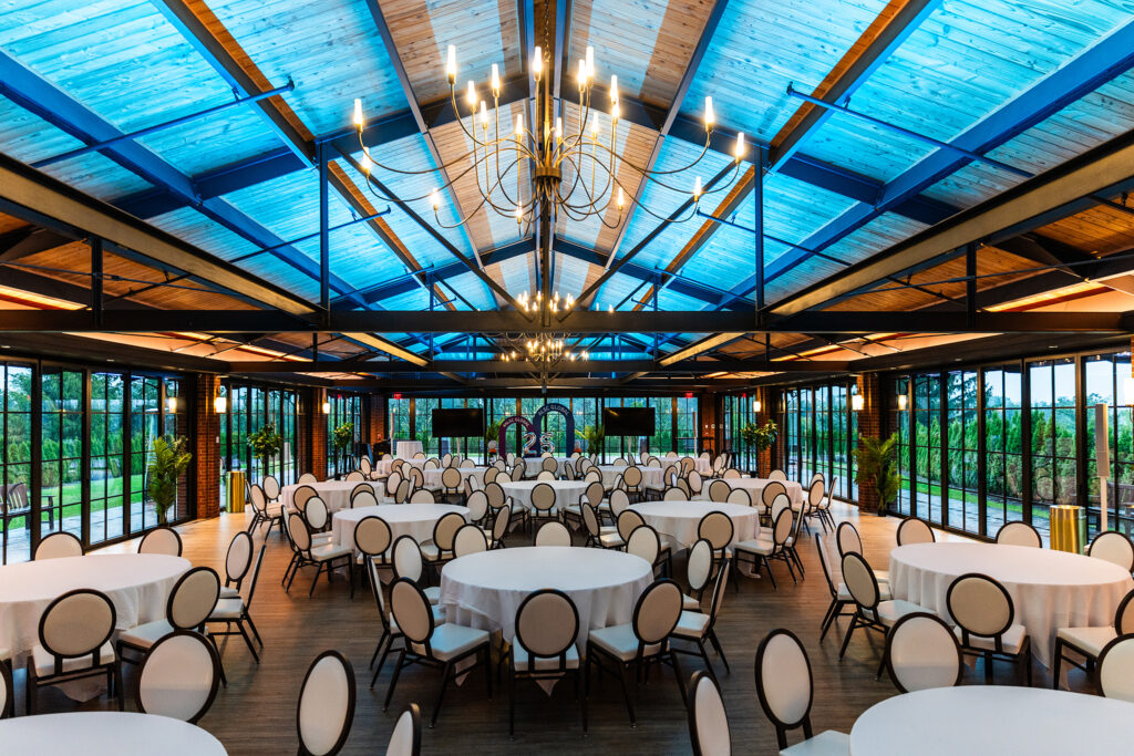 A spacious banquet hall at St Johns Resort near Detroit, with round tables covered in white cloths and surrounded by white chairs, featuring large windows, chandeliers, and a blue glass ceiling letting in natural light for Automotive Logistics and Supply Chain Global events.
