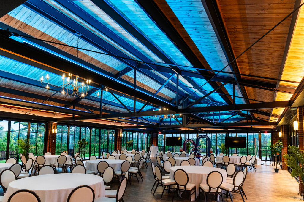 A spacious, elegant banquet hall in Detroit with round tables and white chairs, large glass windows, plants, and a high ceiling with exposed beams and blue-tinted skylights—perfect for corporate events. Warm lighting adds to the inviting atmosphere.