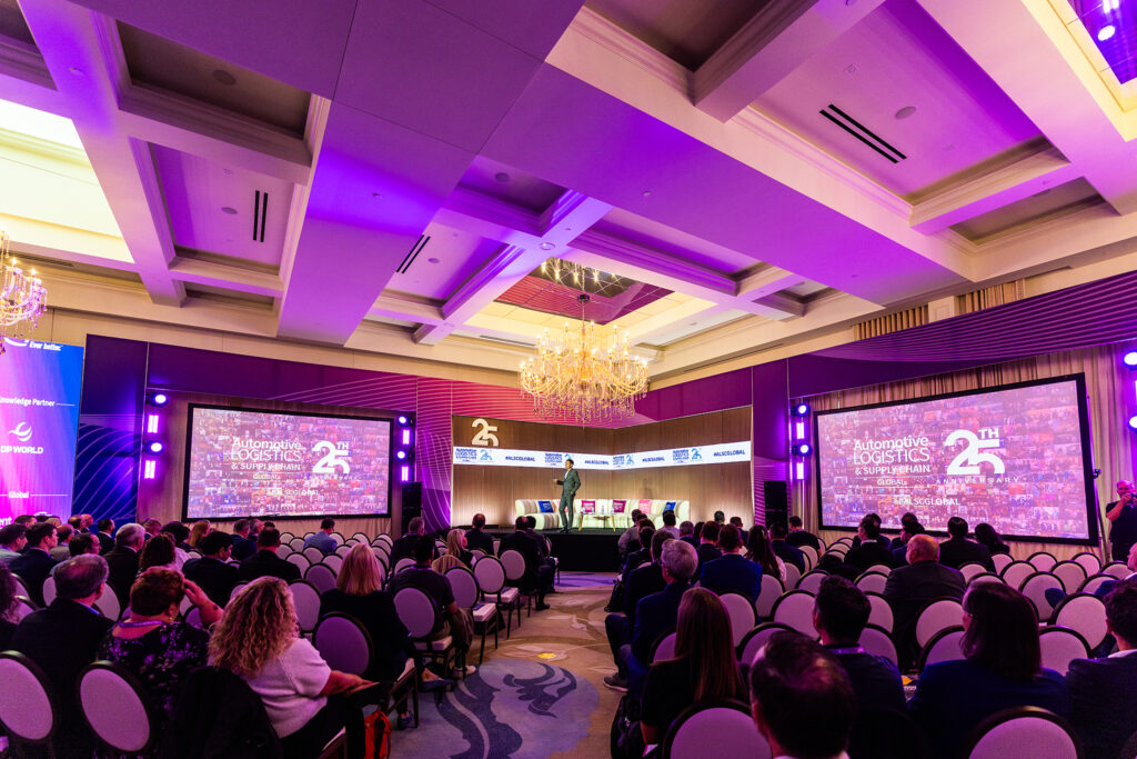 An audience sits in a large, elegant conference room with chandeliers at St Johns Resort in Detroit, watching a speaker on stage at the 25th Automotive Logistics conference (ALCS). Two large screens display the event branding and information.