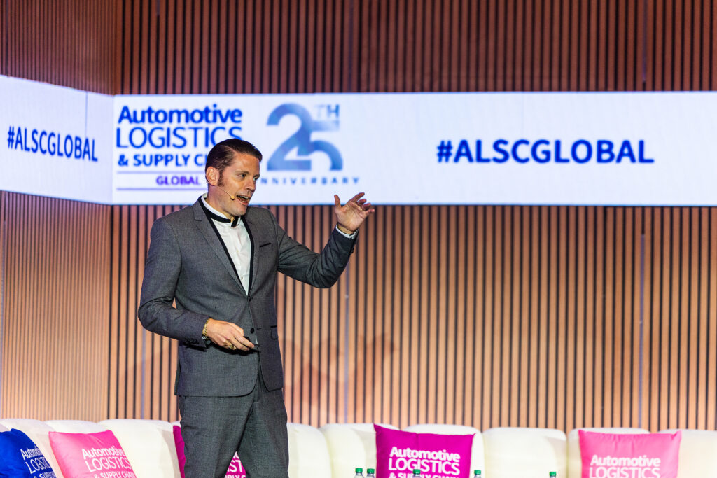 A man in a gray suit speaks on stage at the Automotive Logistics and Supply Chain Global event in Detroit. The backdrop features the ALCS name, 25th anniversary logo, and hashtag #ALSCGLOBAL.