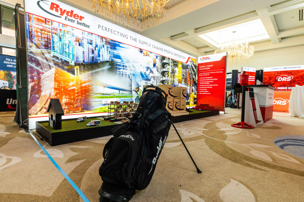 A black golf bag and clubs stand in front of a Ryder Automotive Logistics and Supply Chain Global (ALCS) trade show booth in Detroit, featuring supply chain graphics, a mini putting green, and display screens in a well-lit, carpeted room.