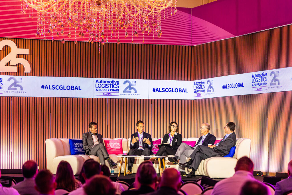 Five people in corporate business attire sit on stage in a panel discussion at the Automotive Logistics and Supply Chain Global (ALCS) conference. A chandelier hangs above, event branding appears on digital screens, and audience members are seated in front.