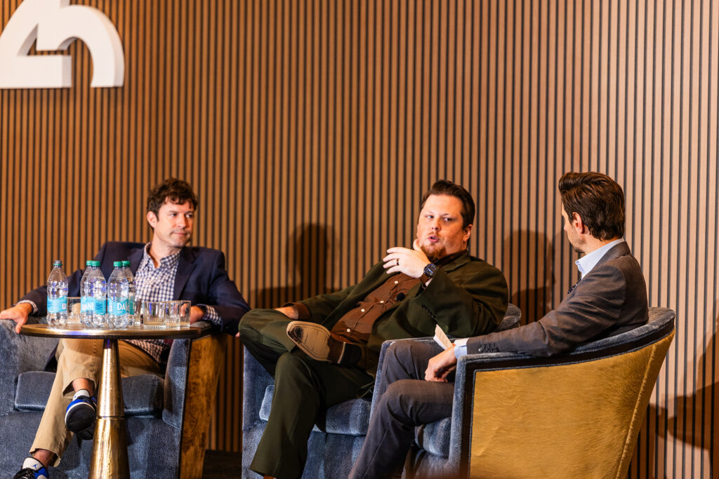 Three men in suits converse on stage at the Automotive Logistics and Supply Chain Global (ALCS) event. Seated in armchairs beside a table with water bottles, they speak in front of a wooden panel wall and a partial logo at St Johns Resort.