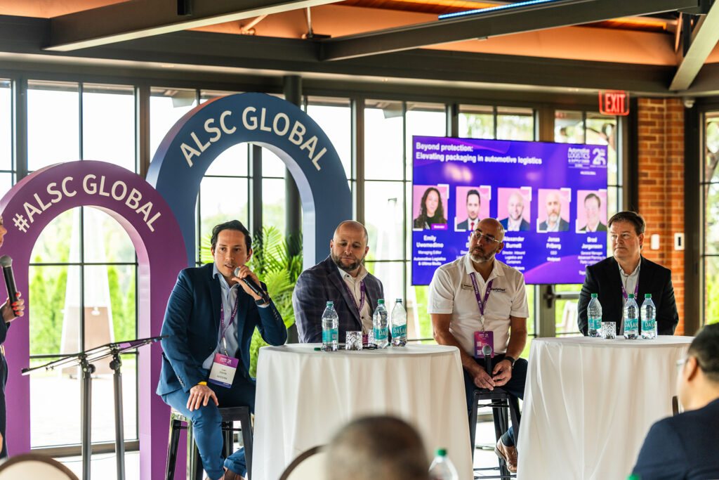 Four men sit at round tables on a panel discussion at the Automotive Logistics and Supply Chain Global event at St Johns Resort. One speaks into a microphone while others listen, with a corporate presentation displayed behind them and bottled water on the tables.