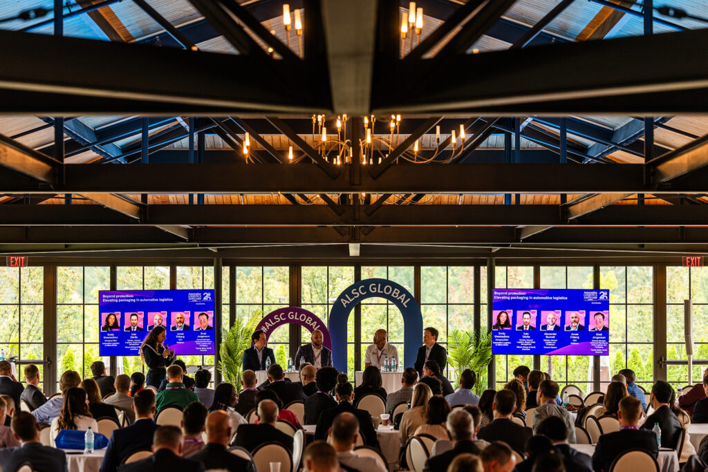 A large audience attends a panel discussion at the Automotive Logistics and Supply Chain Global conference in Detroit, with four speakers on stage under an ALCS GLOBAL sign. Two large screens display speaker information on either side of the stage.